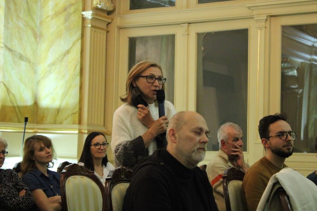 Publiczność siedząca na krzesłach, jedna kobieta z mikrofonem stoi i mówi