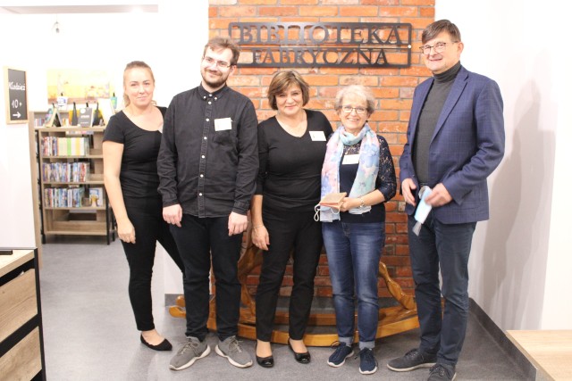 Pięć osób stoi na tle ściany z cegieł i napisu Biblioteka Fabryczna